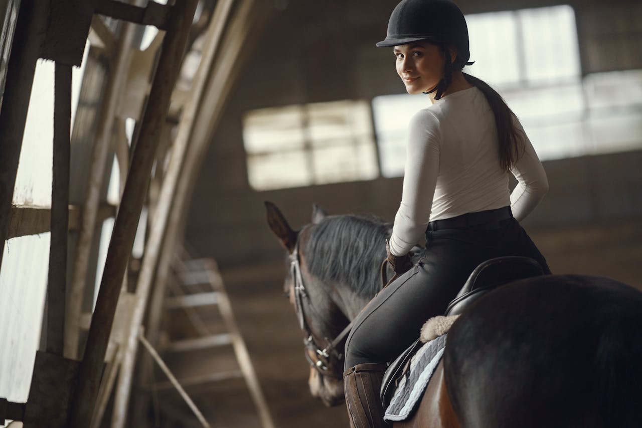 A woman elegantly rides a horse indoors in an equestrian setting, looking back with a smile.
