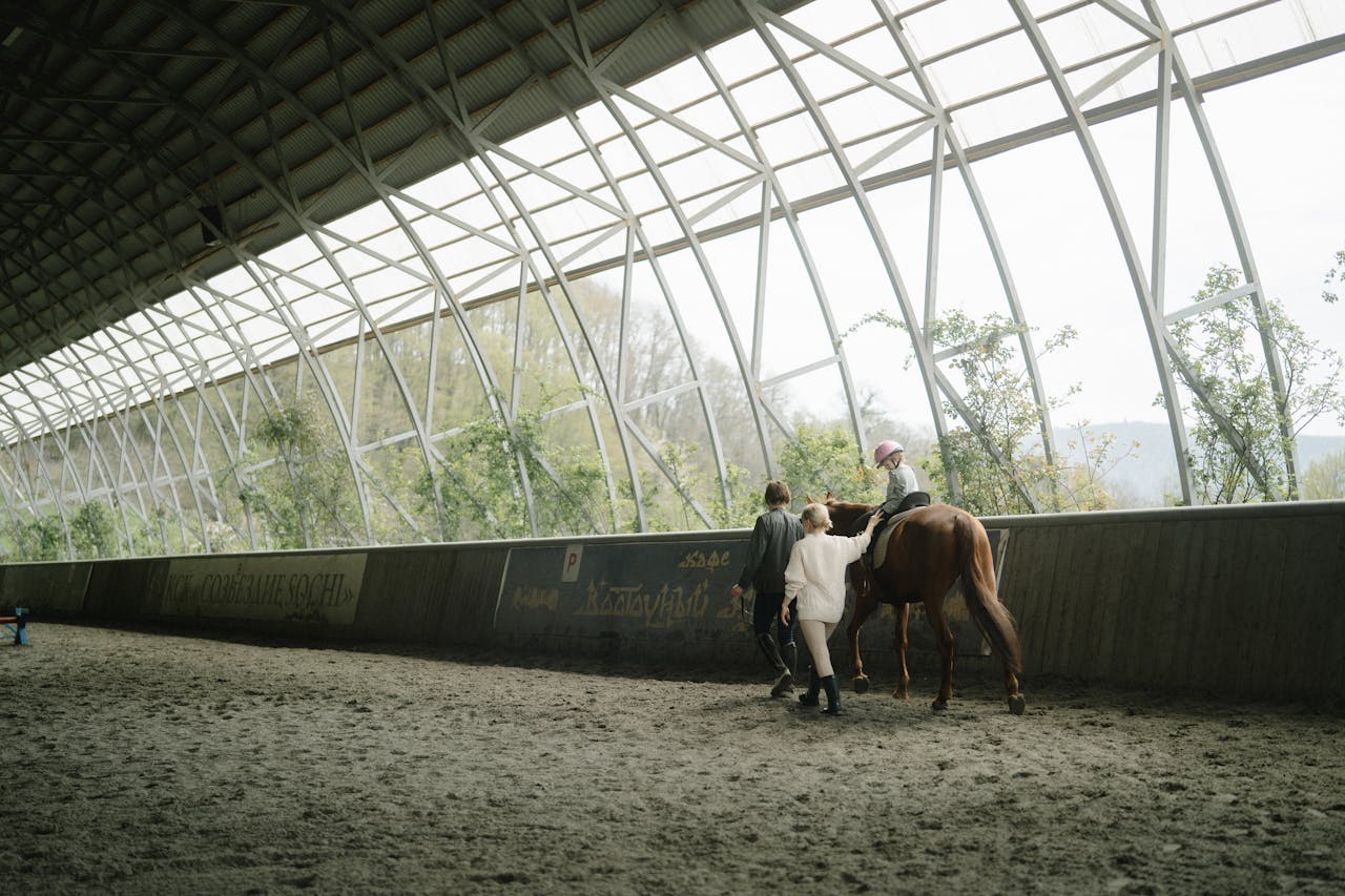 heros-img Child learning horseback riding in an indoor equestrian arena with guidance from instructors.